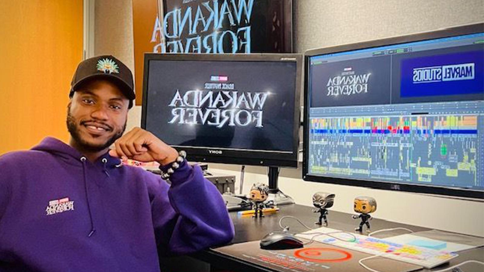 阿兰坐在一张桌子前，桌子上有多个显示器，上面写着“永远的瓦坎达”和“漫威工作室”.” He is wearing a purple hoodie.