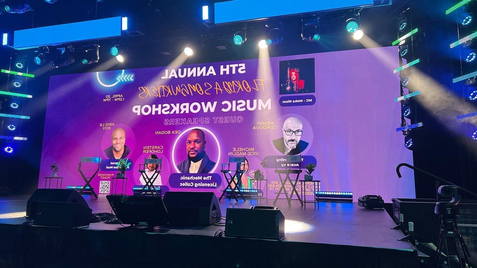 紫色背景的大屏幕上写着“第五届年度词曲作者音乐研讨会”. 屏幕位于灯光明亮的空舞台上方.