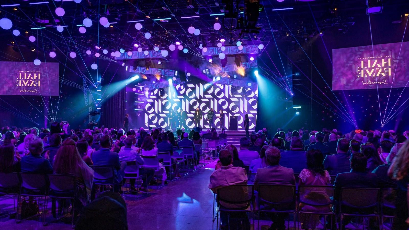 2023年名人堂入选者站在舞台上, 在一个大礼堂里被美丽的紫色灯光包围着. 观众在鼓掌.