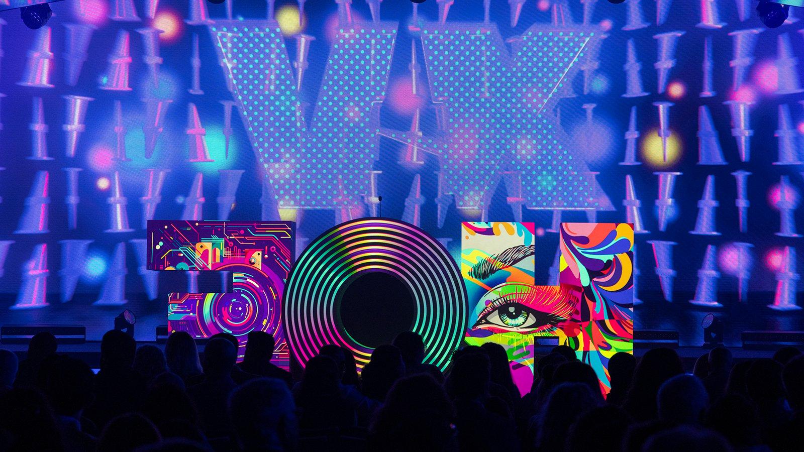 一个巨大的字母“HOF”雕塑坐落在满帆 Live Venue舞台上，每个字母上都投射着不同的彩色图像. 这些字母在一个紫色的大屏幕前，屏幕上用大字体显示着罗马数字“XV”. In front of the stage is a black silhouette of the crowd.