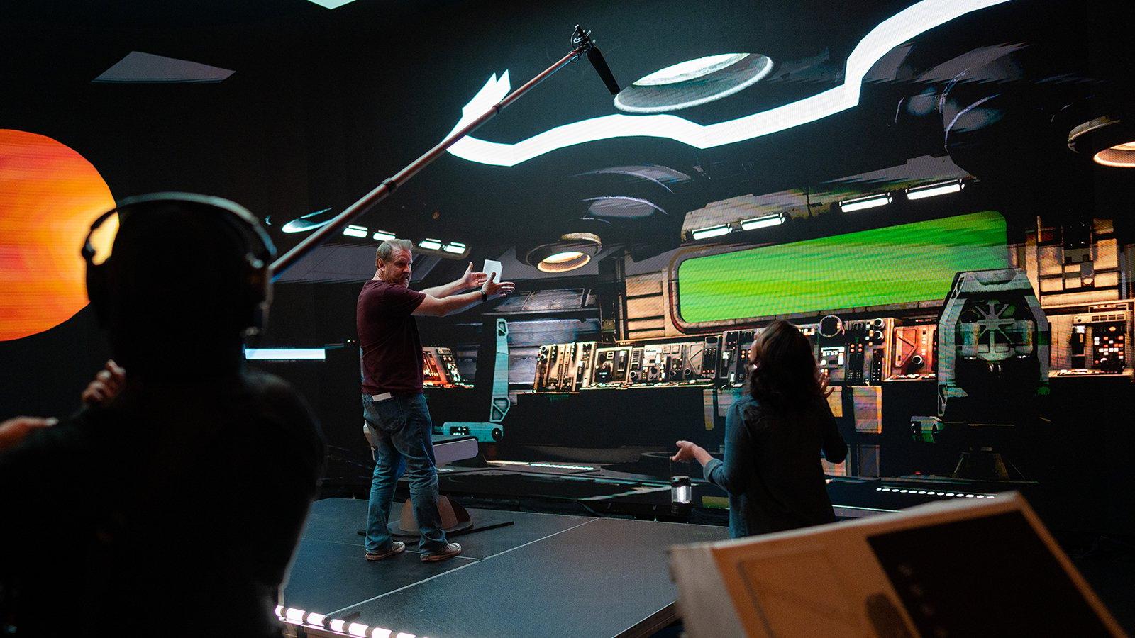空间幼崽’ director Jason Murphy stands onstage in a virtual spaceship set. He is talking to a crew member while another crew member holds a boom mic.