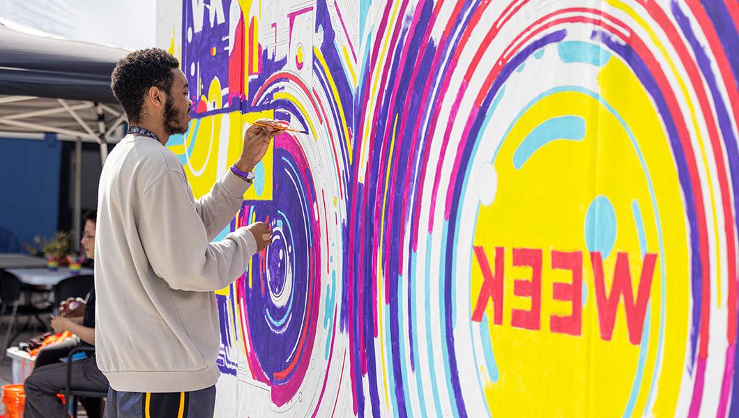 A student wearing a gray sweater paints a purple line on a large mural.