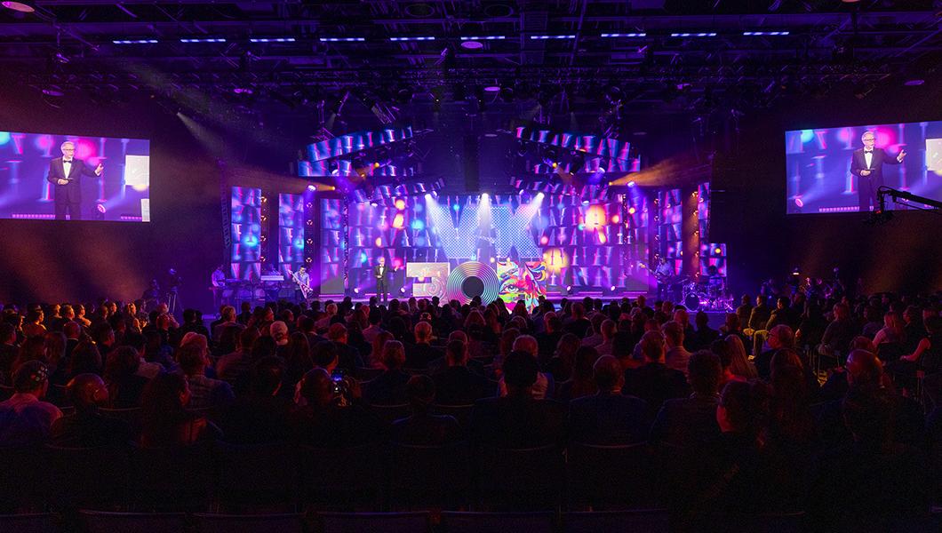A large crowd is seated in front of a raised stage. 在紫色的屏幕前，一个巨大的彩色字母“HOF”雕塑坐落在舞台顶端. 一名身穿黑色西装的男子站在舞台上，并在舞台旁边的屏幕上拍照.